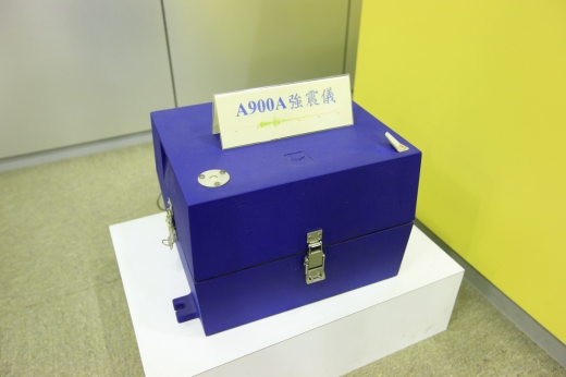 這是一張特寫強震儀的圖。強震儀是一個藍色的箱子，放在一個白色的平台上，箱子有上鎖，其上面有放著此台強震儀型號的立牌。